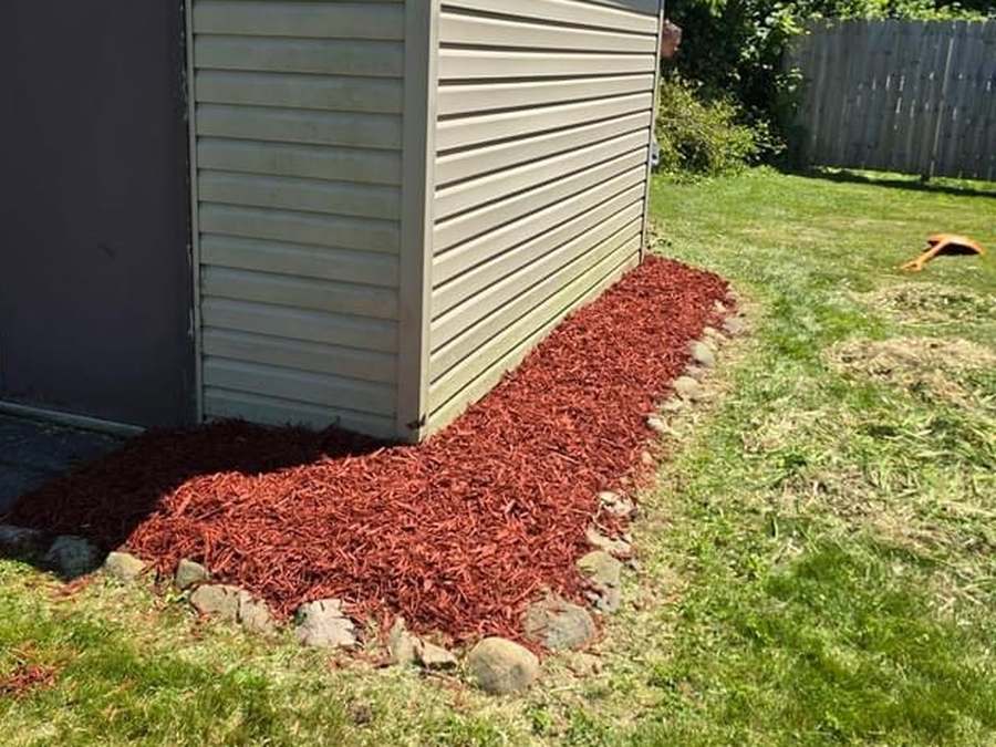 The same shed corner with the bed cleared and covered in fresh red mulch inside the stone edging.