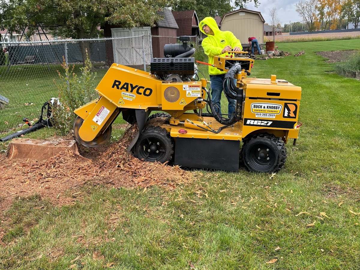 A yellow Rayco stump grinder cutting into a large stump on a lawn while a crew member in a hi-vis hoodie runs it, wood chips piled around the stump.